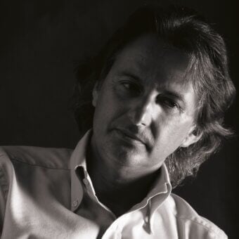 Black and white portrait of Danilo Bonfanti with wavy hair, wearing a light button-down shirt, angled upwards with contemplative gaze, dramatic lighting.