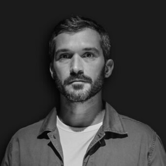 Portrait of a man, Gio Tirotto, with short dark hair and gray facial hair, wearing a light gray button-down shirt against a dark gray backdrop.