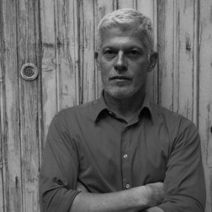 Black and white portrait of a man with short hair, dark shirt, arms crossed, against a weathered wood wall with a metal fixture.