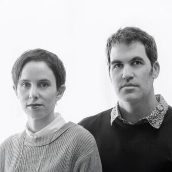 Black and white portrait of a woman in cream and gray knitwear, and a man in a black sweater with patterned shirt, against a white backdrop.