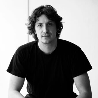 Marco Lavit portrait, black and white photograph, fair skin, wavy dark hair, black crew-neck shirt, crossed arms, plain background.