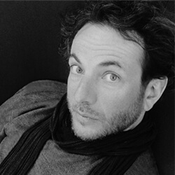 Black and white portrait of Matteo Origoni wearing a dark scarf and light sweater, with short beard and contemplative gaze against a dark background.