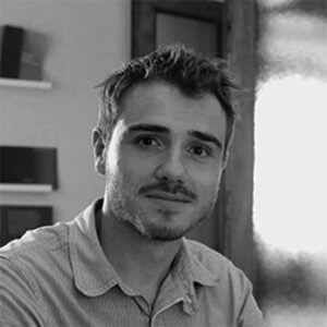 Black and white portrait of Matteo Silverio, wearing a striped collared shirt, with blurred interior background and wooden doorframe.