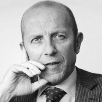 Bald man in pinstripe suit holds vintage mobile phone, serious expression, geometric tie, light gray background, even lighting.