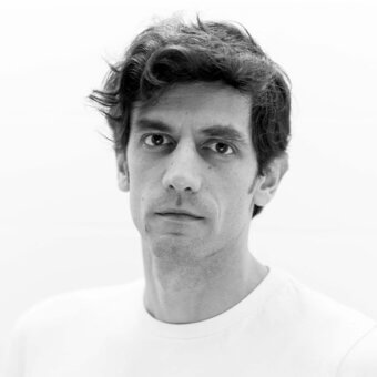 Black and white portrait of Tommaso Caldera, direct gaze, dark wavy hair, white t-shirt, even lighting, close-up headshot, neutral background.