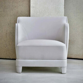 Lounge chair, light grey leather, boxy silhouette with curved backrest and cylindrical legs, set against textured beige and silver wall panels.