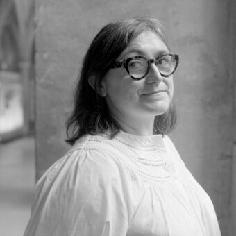Black and white portrait of Ionna Vautrin wearing a white ruffled blouse and tortoiseshell glasses, against a textured neutral wall.