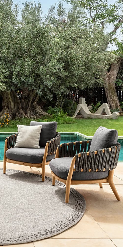 Rope armchairs with curved wood frames and gray cushions sit on a circular rug by a pool, with a lawn and sculpture backdrop.