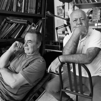 Black and white portrait of two men in a cluttered studio, featuring a chaise lounge, spindle chair, bookshelves, scanner, and printer.