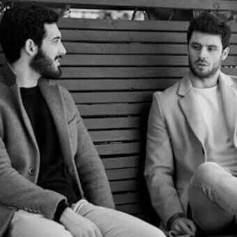 Black and white photograph of two men on a wooden bench, wearing blazers and light trousers, with a slatted back creating linear patterns.
