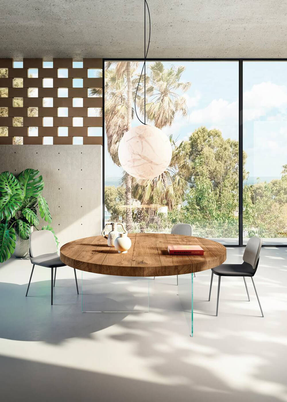 Round dining table with light-brown wood top and glass base, surrounded by four dark gray chairs in a modern room with concrete walls.