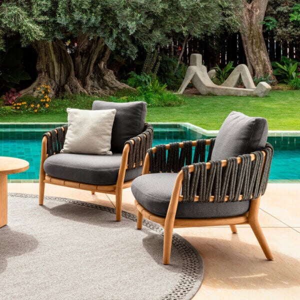Outdoor armchairs with wood frames and woven rope detailing sit on a gray rug by a turquoise pool, featuring dark gray cushions.