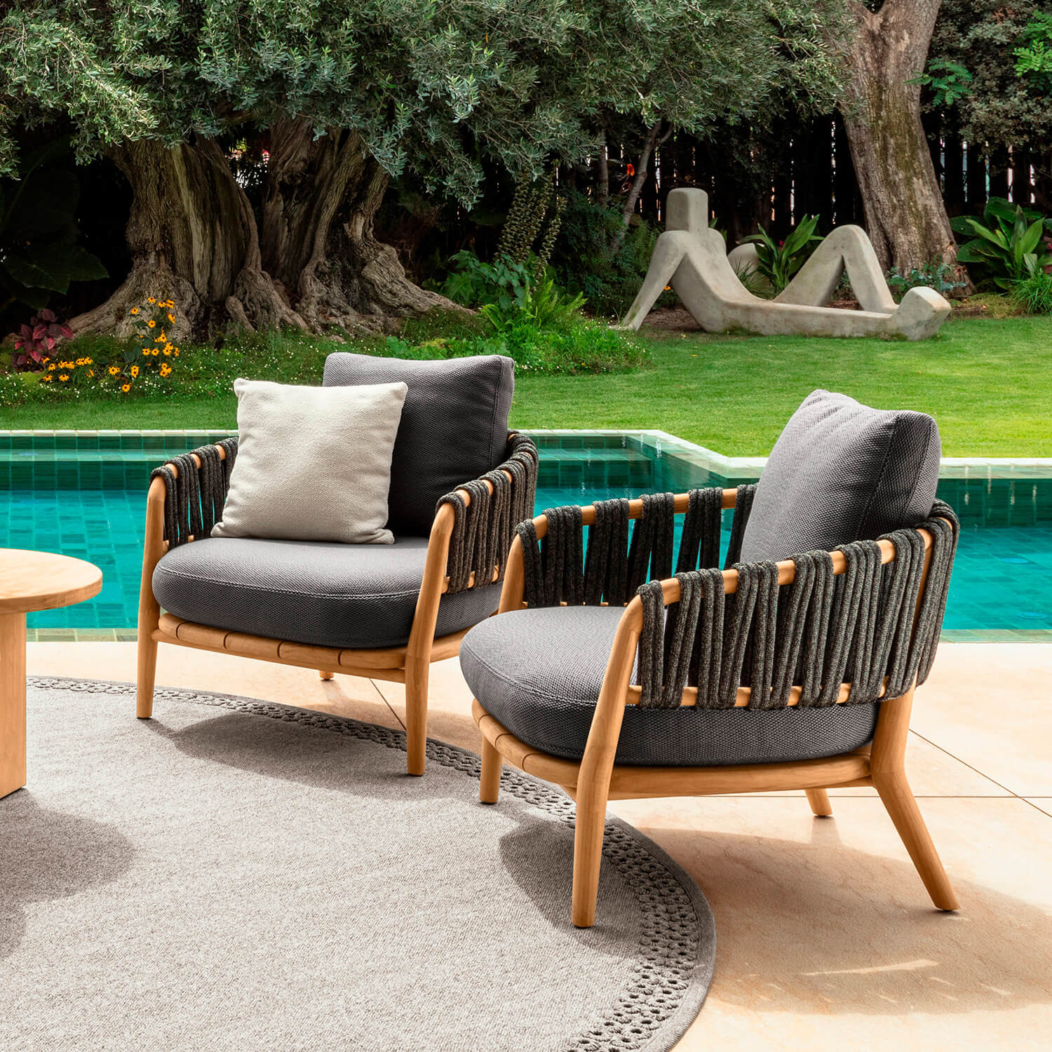 Outdoor armchairs with wood frames and woven rope detailing sit on a gray rug by a turquoise pool, featuring dark gray cushions.
