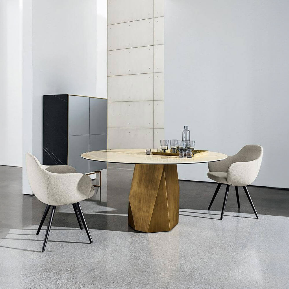 Marble round table with brass faceted base, flanked by beige upholstered chairs in a minimalist interior with gray concrete flooring.
