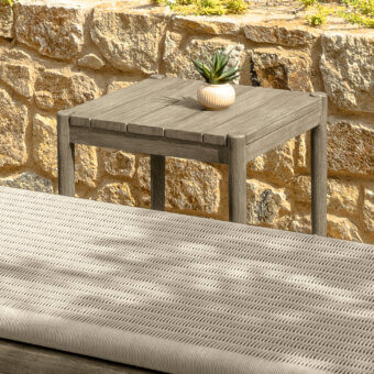 Talenti Dolcevita side table, weathered gray finish, square shape, with succulent; adjacent woven gray chaise lounge, stone wall backdrop.