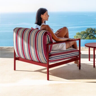 Talenti Nalu lounge chair, red aluminum frame, striped cushions, positioned on a beige tile terrace with ocean and hillside view.