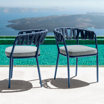 Talenti Swipe armchairs, dark blue metal frame, rope backrests, gray cushions on tiled terrace overlooking turquoise pool and caldera view.