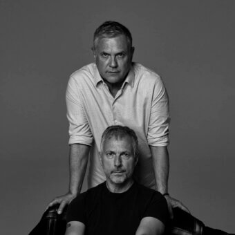 Black and white portrait of two men against gray, one with gray hair in a light shirt, the other bearded in black, studio lighting.