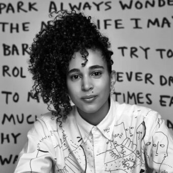 Black and white portrait of Shantell Martin in illustrated white shirt, with handwritten text wall backdrop and soft, even studio lighting.