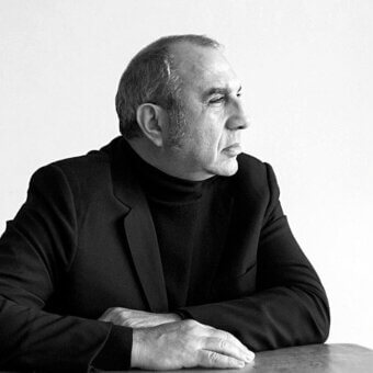 Stefano Giovannoni portrait, grayscale photograph, man with gray hair wearing a black turtleneck and blazer, seated with crossed arms.