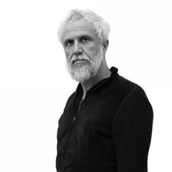 Man portrait, grey hair and beard, dark textured jacket, neutral expression, white background, diffused lighting, studio nucleo promotional image.