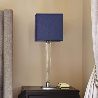 Spillo table lamp, chrome and navy blue, features a cylindrical glass base with a square fabric lampshade on a dark wood table.