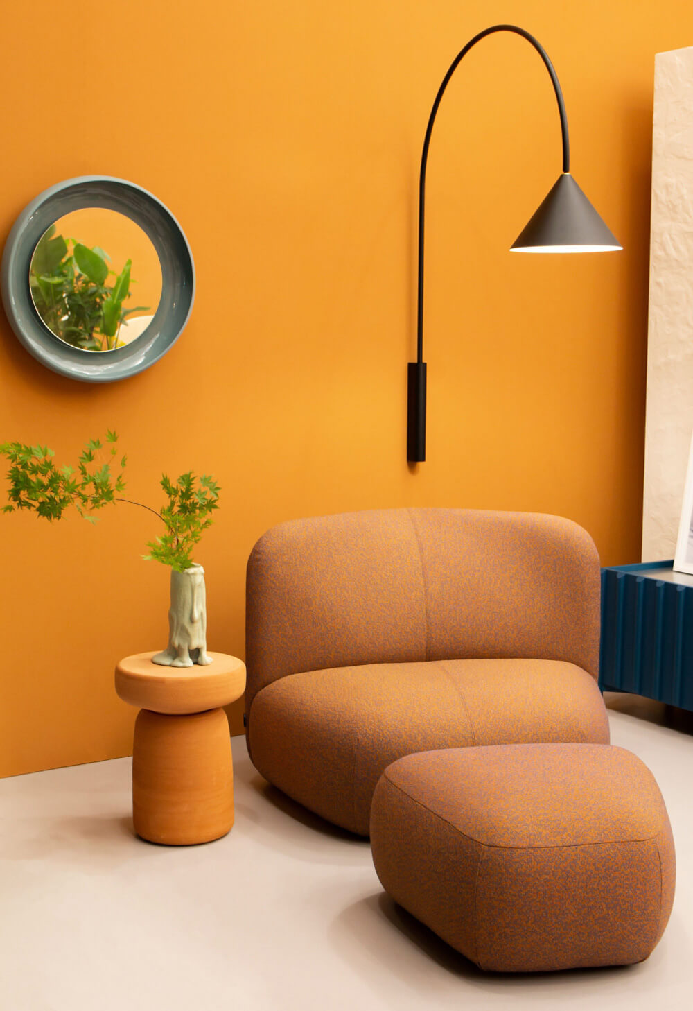 Lounge chair and ottoman upholstered in rust-orange fabric, rounded organic shape, with terracotta side table and teal mirror.