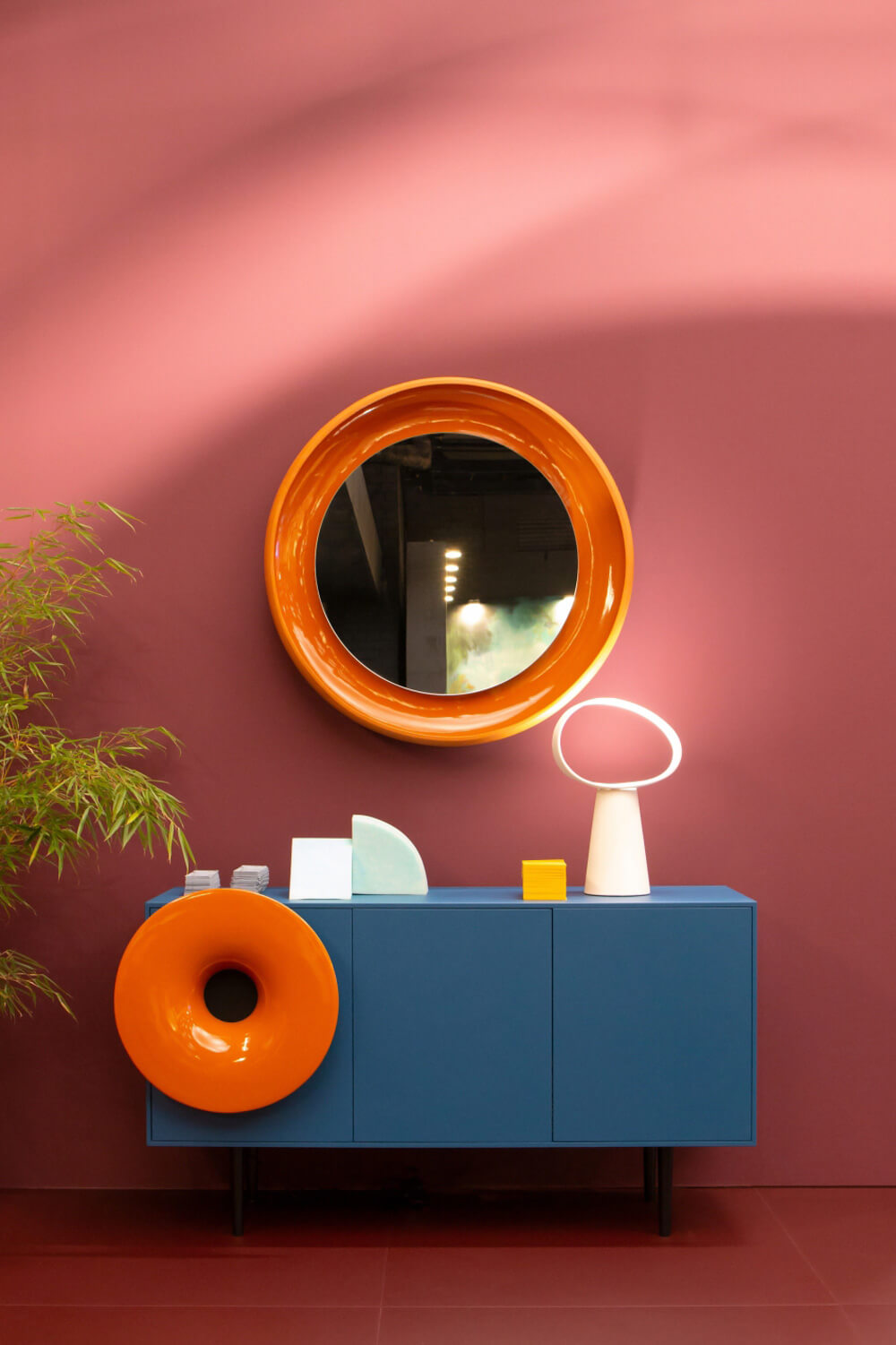 Miniforms Caruso bar cabinet, deep blue, circular mirror; orange donut, ceramic vases, plant on reddish-brown tile floor against dusty rose wall.
