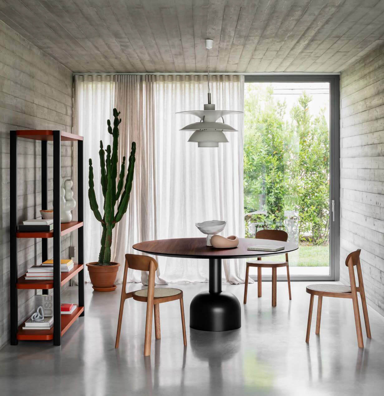 Round illo table with walnut top and gray base, surrounded by four light wood chairs in a concrete room with shelving.