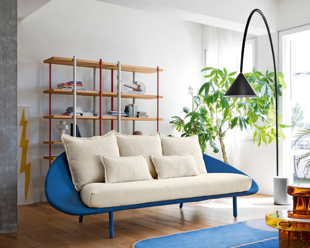 Lem sofa, light beige fabric, curved two-seater with blue base and armrests, paired with red shelving, black lamp, and yellow graphic pillar.
