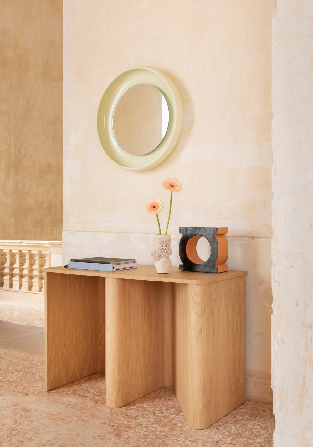 Nami console in light wood with curved form, pale green mirror above, vase with gerbera, book, and geometric sculpture on terrazzo floor.