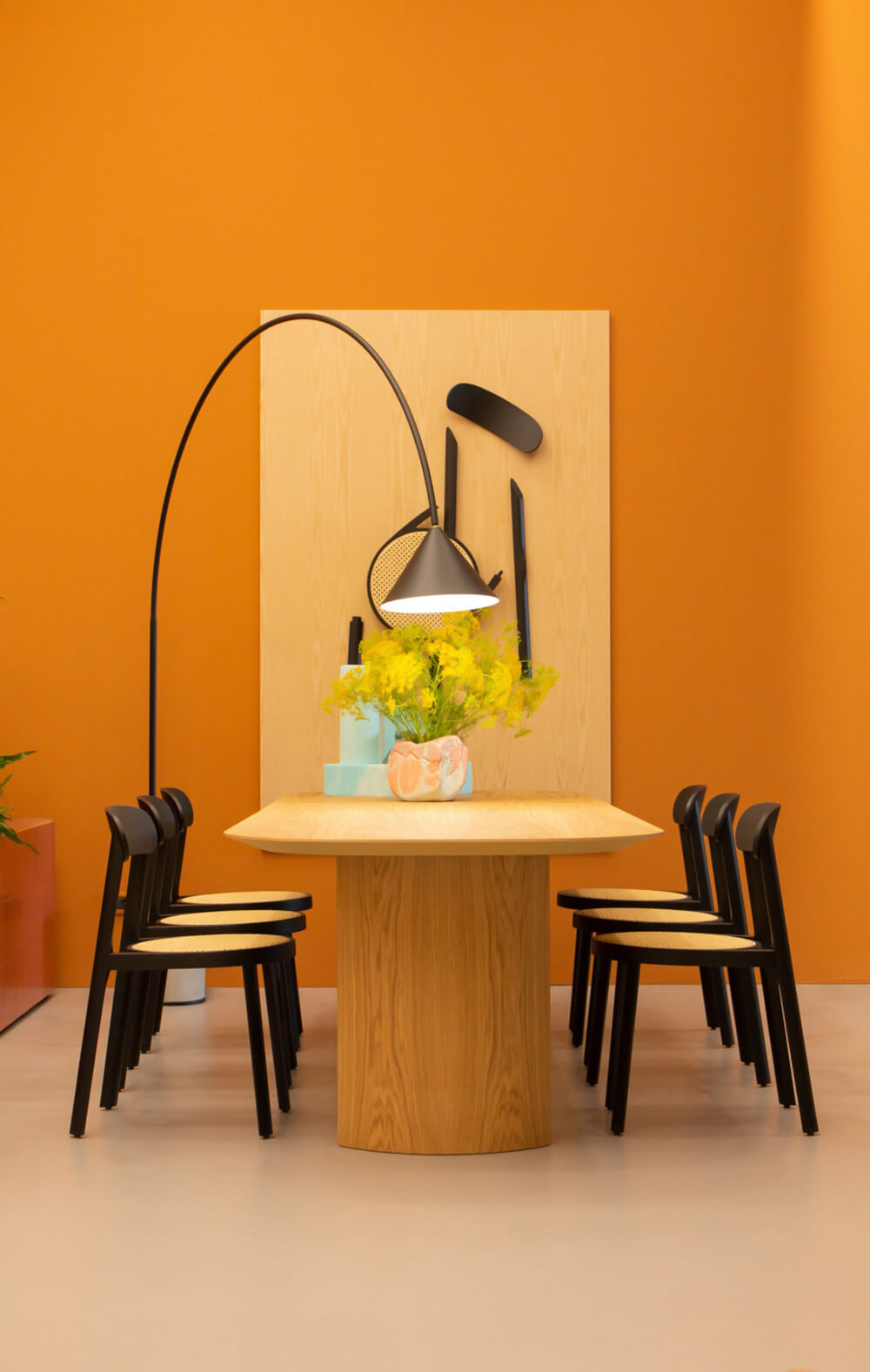 Miniforms Plauto table, light wood top, cylindrical base, with four black chairs, orange wall, gray floor, and arched floor lamp.