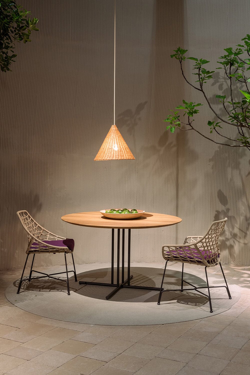 Round wood table with black metal base, green bowl, and woven chairs, under rattan pendant light in minimalist outdoor setting.