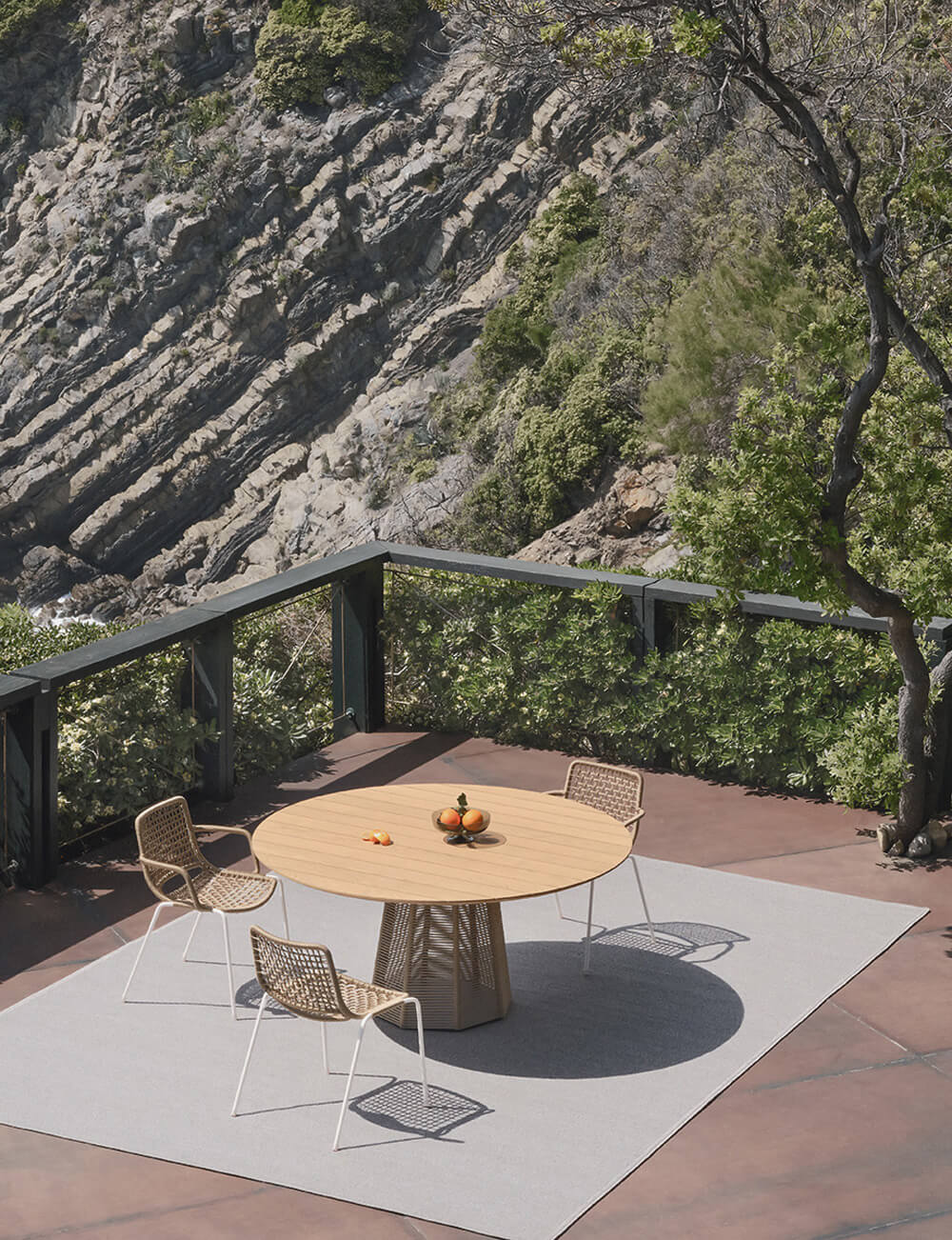Potocco Sophie table, wood top, woven base, on gray rug with four woven chairs, oranges, terracotta patio, metal railing, foliage backdrop.