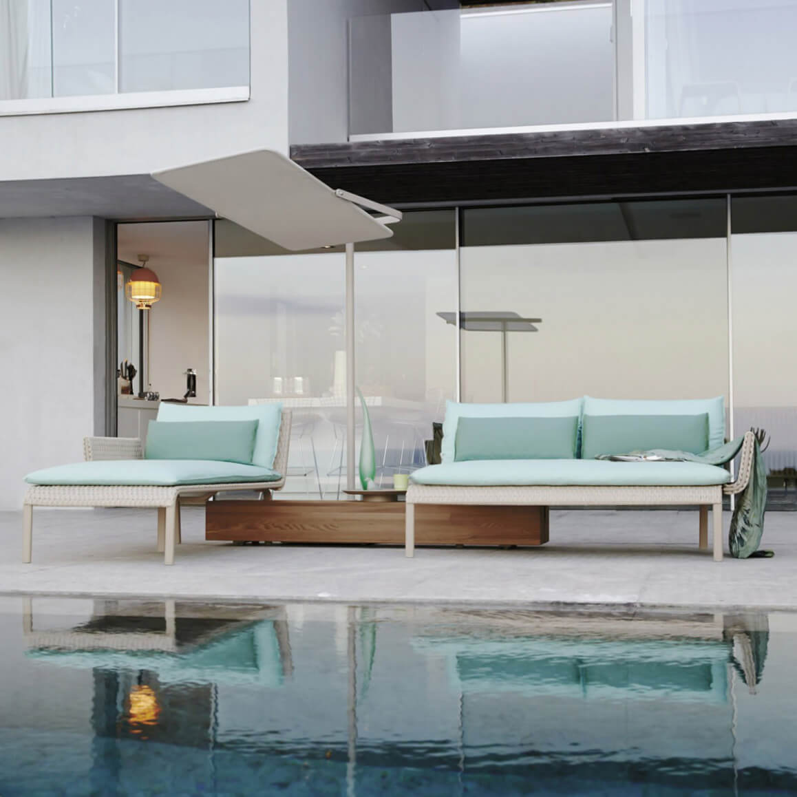 Rattan lounger with turquoise cushions on concrete patio by pool, beige umbrella, modern building backdrop, light neutral palette.