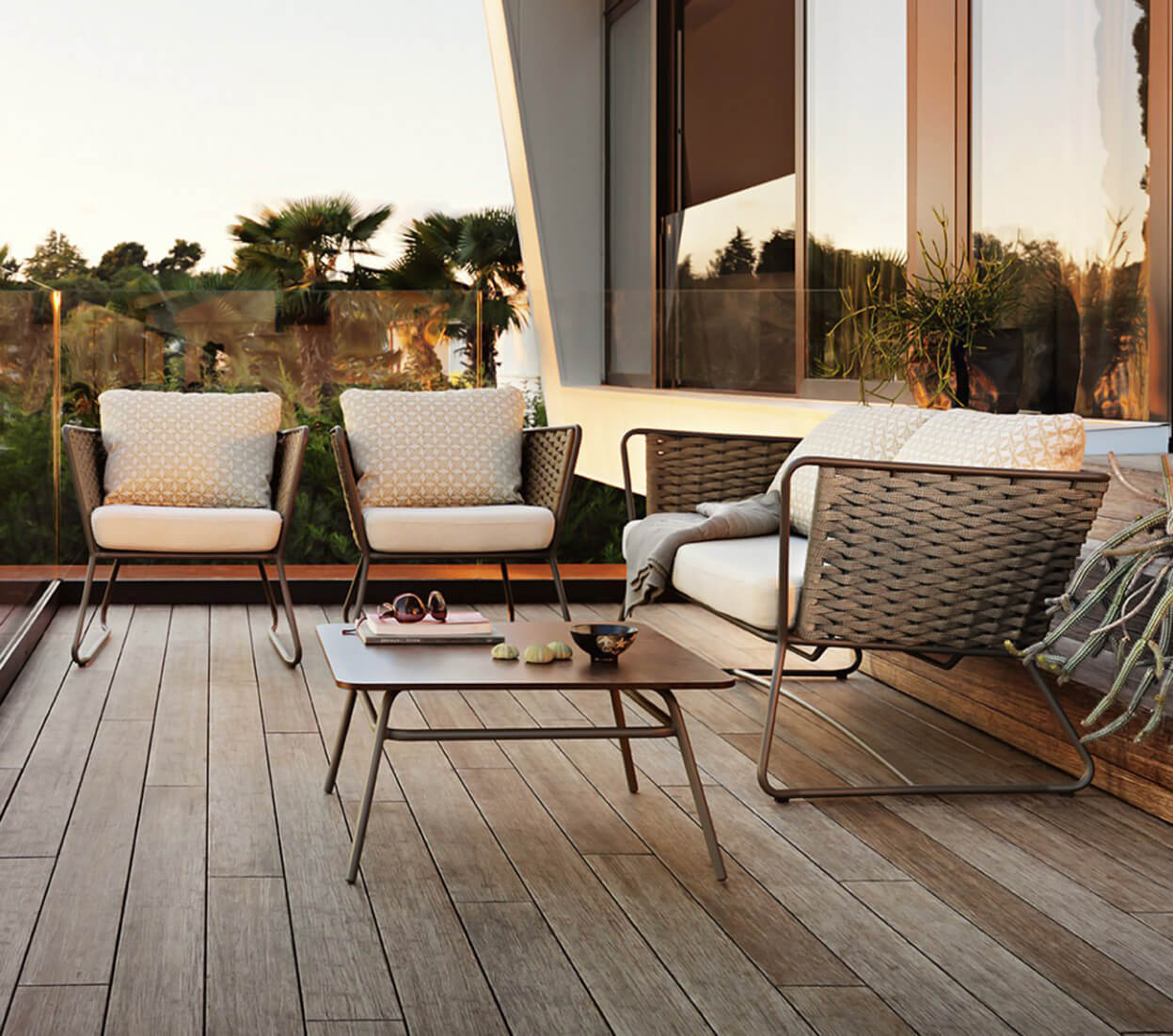 Rattan outdoor loveseat and armchairs with cream cushions, dark gray metal frames, and a bronze coffee table on a wood deck.
