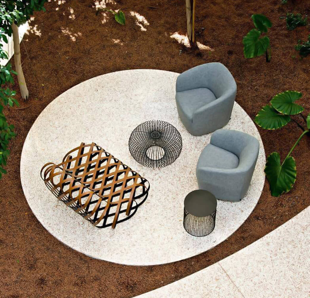 Outdoor lounge chairs, blue-gray fabric, curved armrests on speckled stone patio with woven basket, metal side table, and lush green foliage.