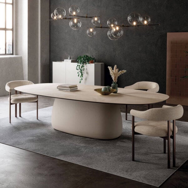 Oval dining table with beige textured top, curved base, and four beige fabric chairs with dark metal legs, under a glass orb chandelier.