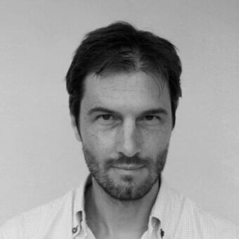 Black and white portrait of Marco Gregori, featuring dark hair, a trimmed beard, and a light patterned shirt, with direct eye contact.