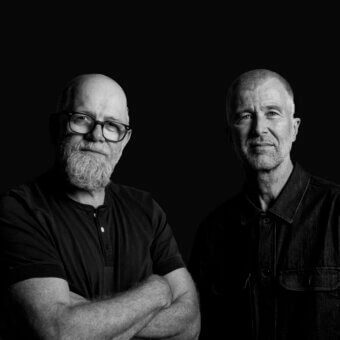 Black and white portrait of two men with gray beards against a black background, wearing dark shirts with neutral expressions.
