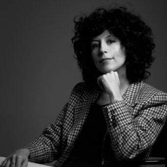 Black and white portrait of Arianna Crosetta wearing a houndstooth blazer, dark top, and curly hair, with a gradient gray background.