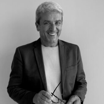 Portrait of a man in a blazer and sweater holding glasses, gray hair, neutral backdrop, studio lighting, euro sironi and Cassoni branding.