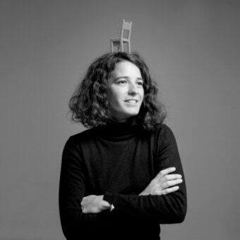 Woman with curly hair balancing a small wooden chair on her head, wearing a black turtleneck, against a gray background.