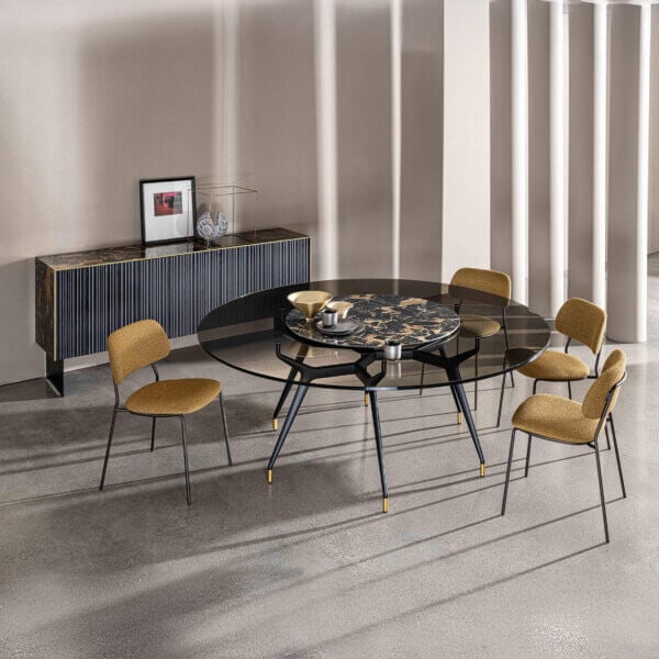 Round glass dining table with black metal base and gold feet, paired with mustard yellow armchairs, in a modern beige and gray interior.