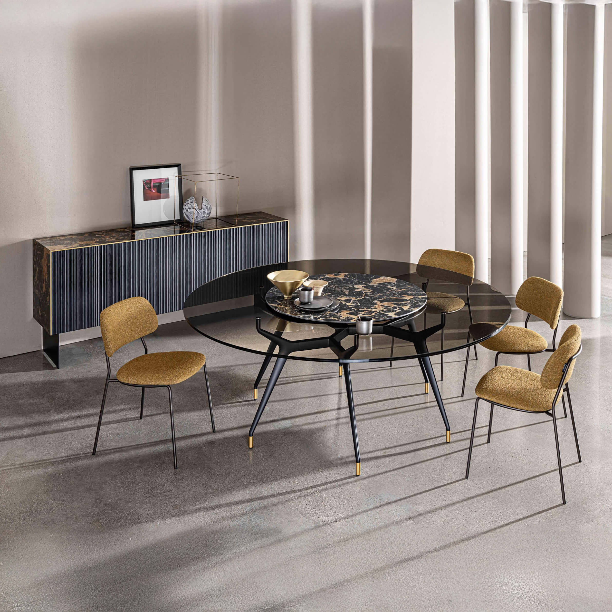 Round glass dining table with black metal base and gold feet, paired with mustard yellow armchairs, in a modern beige and gray interior.
