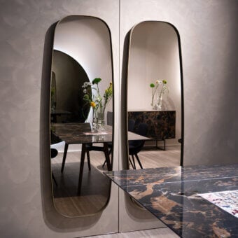 York arched mirrors by Sovet Italia, brushed brass frames, reflect dining area with marble table, black chairs, and floral arrangement.