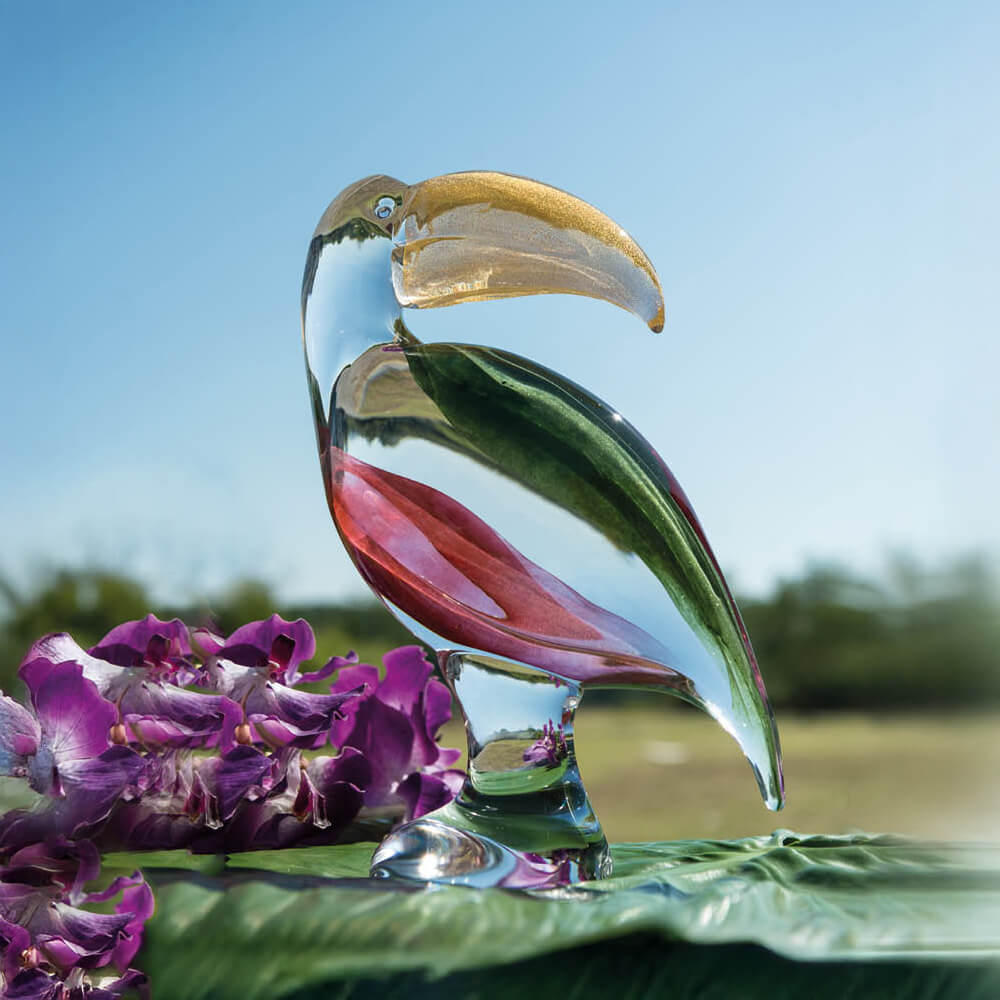 Glass toucan sculpture with green & pink swirls, gold beak, on glossy leaf with purple orchids, clear base, blurred garden backdrop.