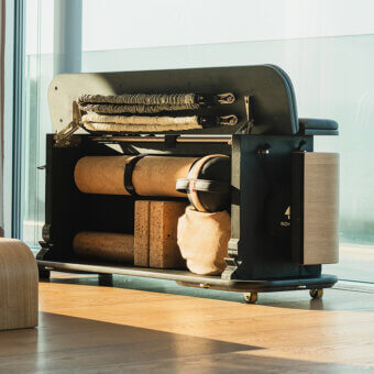 Wellness machine with dark gray metal frame, wooden carriage, and cork cylinders, against blurred city view.