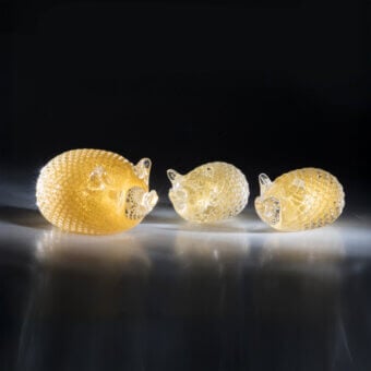 Glass hedgehog sculptures, golden tone, curled pose, arranged in descending size on reflective dark gray surface against black backdrop.