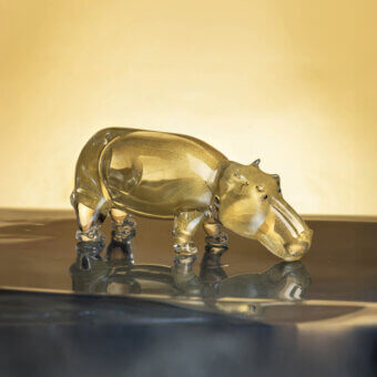 Glass hippopotamus sculpture, golden finish, rounded form on reflective black surface, warm gradient background.